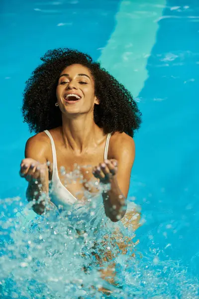 Joyeuse femme noire éclaboussant l'eau à l'intérieur de la piscine, se délectant de son plaisir et de plaisir pendant les vacances — Photo de stock