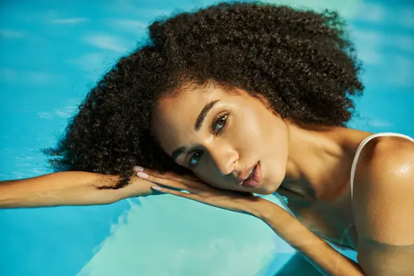 Porträt einer jungen afrikanisch-amerikanischen Frau mit lockigem Haar, die in die Kamera schaut und im Pool schwimmt — Stockfoto