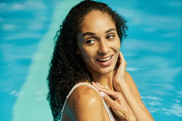 Feliz jovem mulher negra com cabelo encaracolado molhado sorrindo enquanto em pé na água da piscina azul — Fotografia de Stock