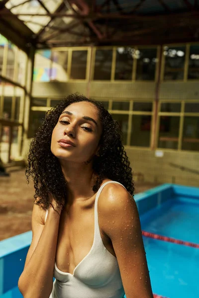 Jeune femme afro-américaine détendue avec les cheveux bouclés humides et la peau debout près de la piscine, sereine — Photo de stock
