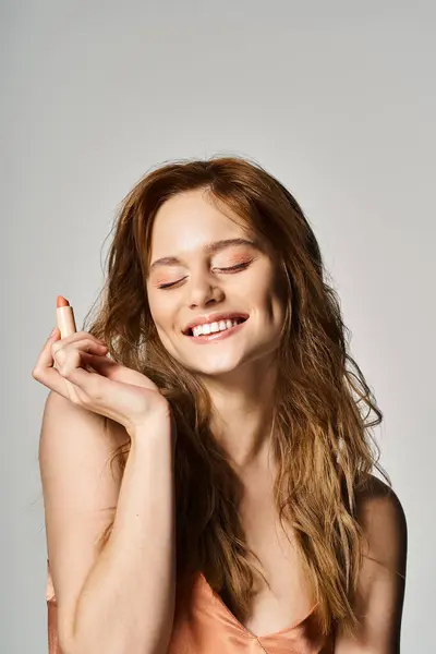 Foto de belleza de la mujer riendo con los ojos cerrados sosteniendo lápiz labial sobre fondo gris. Pelusa de melocotón - foto de stock
