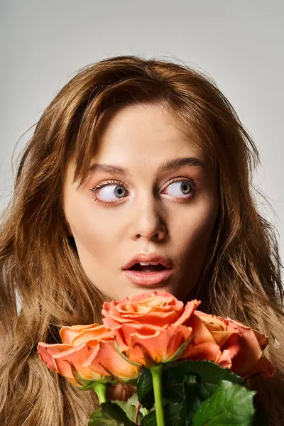 Retrato de mujer sorprendida con ojos azules, sosteniendo rosas melocotonas posando sobre fondo gris - foto de stock