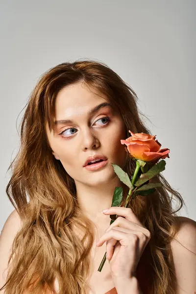 Beauty shot of pretty curious woman with blue eyes, holding peachy rose near face on grey background — Stock Photo