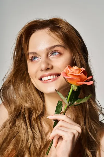 Красивая улыбающаяся молодая женщина с голубыми глазами, с персиковой розой возле лица на сером фоне — стоковое фото