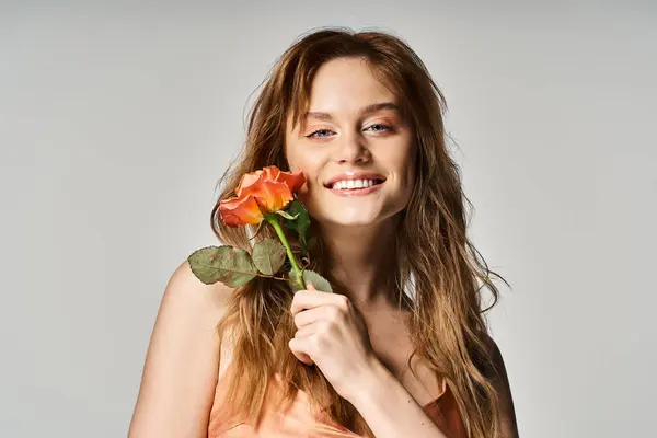 Веселая улыбающаяся привлекательная девушка с голубыми глазами, с персиковой розой возле лица на сером фоне — стоковое фото