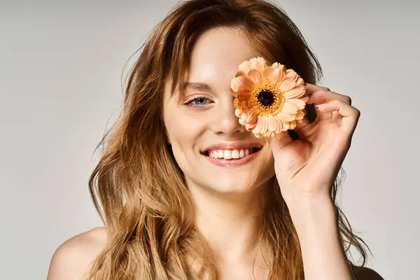 Schöne lächelnde junge Frau mit blauen Augen, mit Gerbera-Gänseblümchen in der Nähe Auge auf grauem Hintergrund — Stockfoto