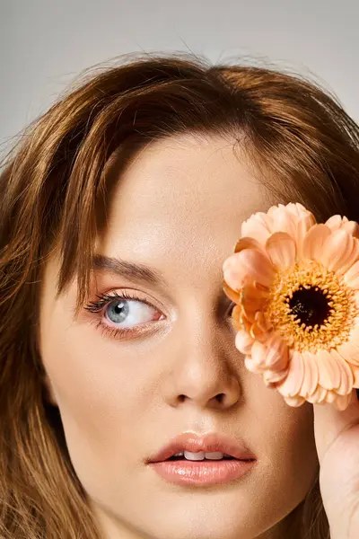 Closeup beauty shot of pretty woman with peach natural makeup and daisy near eye on grey background — Stock Photo