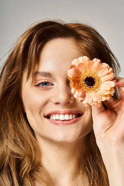 Plan beauté de femme souriante avec maquillage pêche et gerbera marguerite près des yeux sur fond gris — Photo de stock