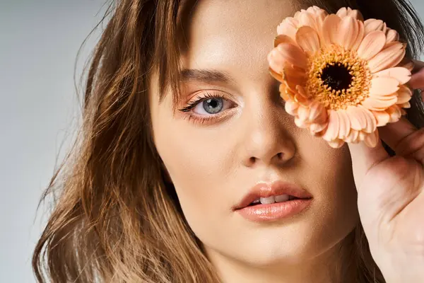 Closeup beauty shot of pretty woman with peach makeup and gerbera daisy near eye on grey background — Stock Photo