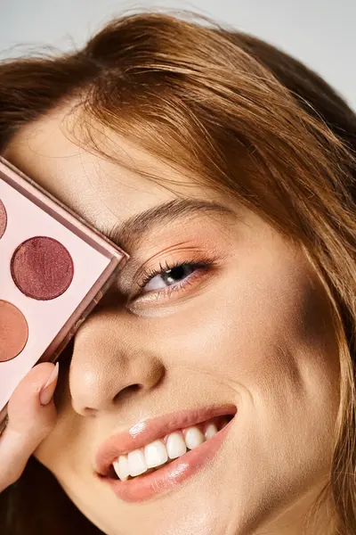 Closeup beauty shot of smiling woman with peach makeup palette near face on grey background — Stock Photo