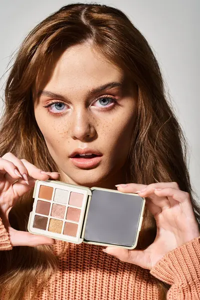 Astonished attractive woman with peach makeup palette near chin on grey studio background — Stock Photo