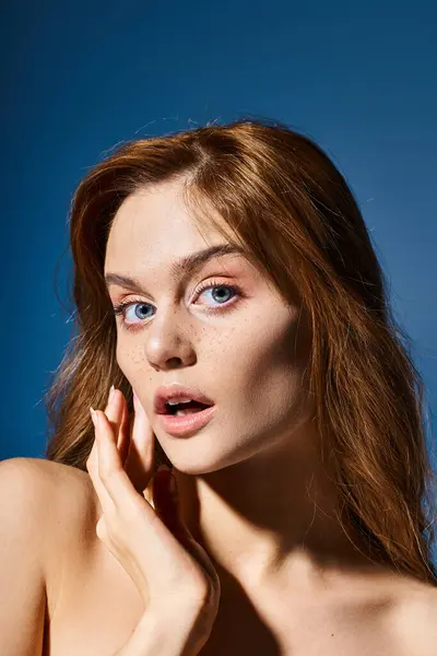 Foto de belleza de mujer asombrada con maquillaje de melocotón y pecas, tocando la mejilla sobre fondo azul - foto de stock