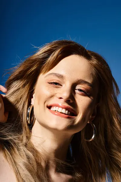Retrato de belleza de una mujer sonriente mirando a la cámara y tocándose el pelo sobre fondo azul - foto de stock