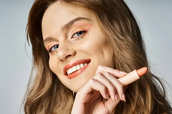 Cheerful woman with face jewels in beige jacket, holding nude lipstick, looking at camera — Stock Photo