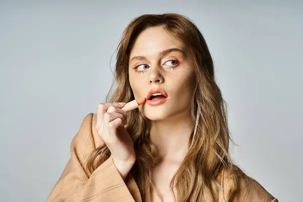 Mujer atractiva con joyas de la cara en chaqueta beige, aplicación de lápiz labial y mirando hacia otro lado, pelusa de melocotón - foto de stock
