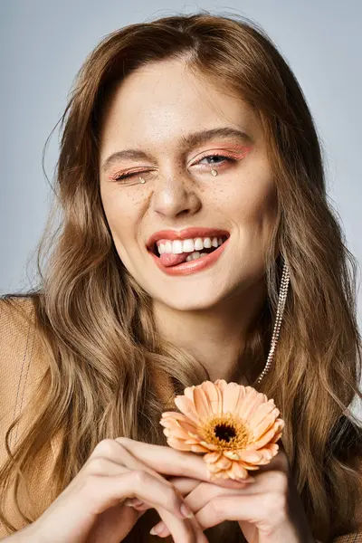 Подмигивая веселой женщине, смотрящей в камеру и держащей маргаритку, носящей персиковый макияж, украшения для лица — стоковое фото