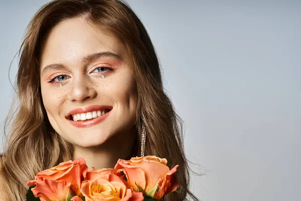 Mulher sorridente olhando para a câmera, segurando rosas e vestindo maquiagem pêssego nu e jóias rosto — Fotografia de Stock