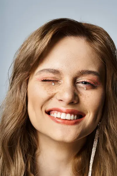 Primer plano de mujer sonriente guiñando con maquillaje de melocotón, joyas de la cara y pecas sobre fondo gris - foto de stock
