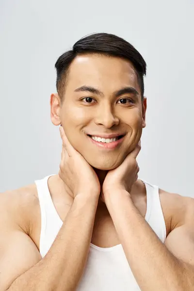 A handsome Asian man with a cheerful smile, hands resting on his chin in a grey studio setting. — Stock Photo