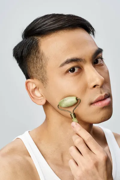 Handsome Asian man using jade roller on his skin in a grey studio. — Stock Photo