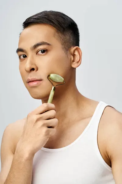 Un hombre asiático guapo está usando suavemente un rodillo de jade como parte de su rutina de cuidado de la piel en un estudio gris. - foto de stock
