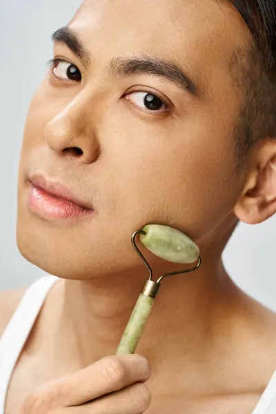 Hombre asiático guapo sosteniendo un rodillo de jade hasta la cara en un estudio gris para una rutina de cuidado de la piel única. - foto de stock