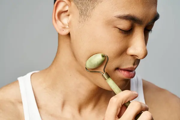 Un hombre asiático guapo en un estudio gris sosteniendo un rodillo de jade - foto de stock