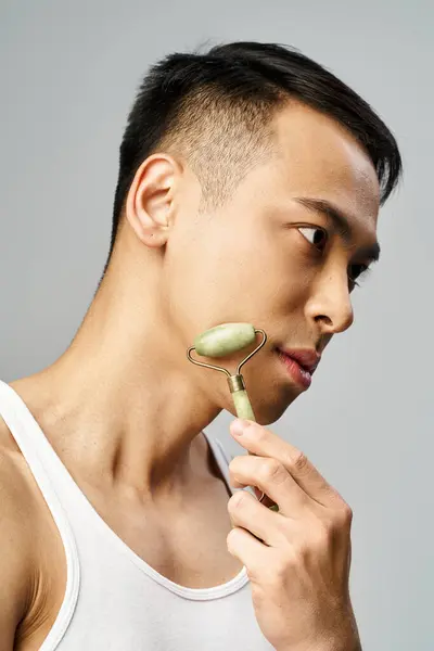 Hombre asiático guapo cuidadosamente usando rodillo de jade en un moderno estudio gris. - foto de stock