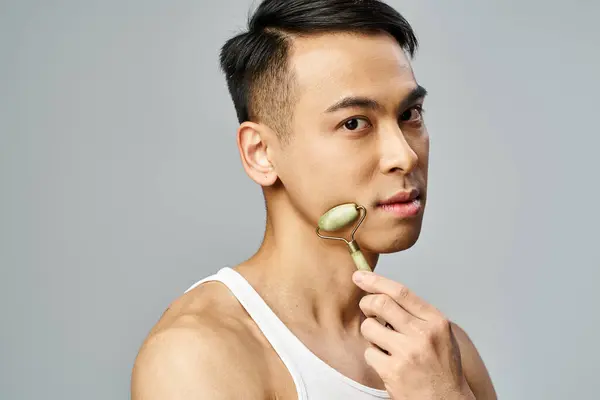 Asian man using jade roller, focusing on grooming and self-care routine in a grey studio setting. — Stock Photo