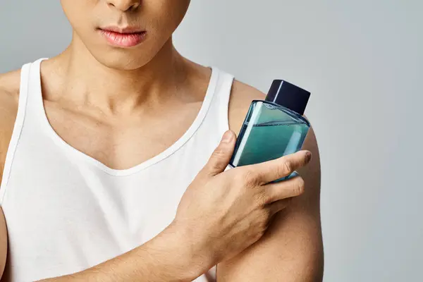 Un Asiatique suave en débardeur tient une bouteille de parfum d'une manière sophistiquée dans un décor de studio gris. — Photo de stock