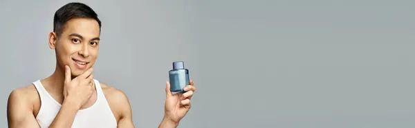 Ein gut aussehender asiatischer Mann hält eine Flasche Parfüm in einem grauen Studio-Ambiente, Banner — Stockfoto
