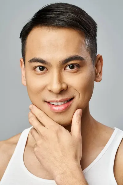 Schöner asiatischer Mann, in einem Tank-Top, posiert selbstbewusst in einem grauen Studio während einer Hautpflege-Routine. — Stockfoto