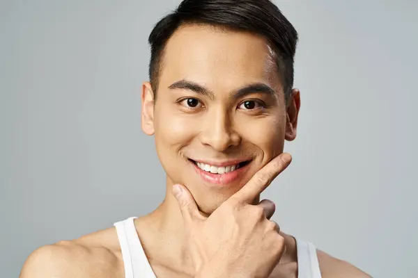 Un hombre asiático guapo con una sonrisa en la cara, exudando felicidad y confianza en un estudio gris mientras usa productos para el cuidado de la piel. - foto de stock