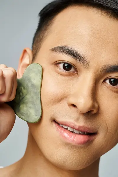 Handsome Asian man in grey studio holding gua sha to face for skincare routine. — Stock Photo