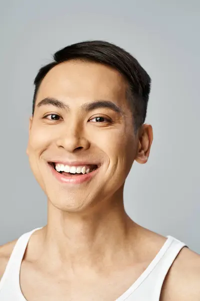 Un hombre asiático guapo está radiante con una sonrisa, exudando calidez y felicidad en un estudio gris mientras usa productos para el cuidado de la piel. - foto de stock