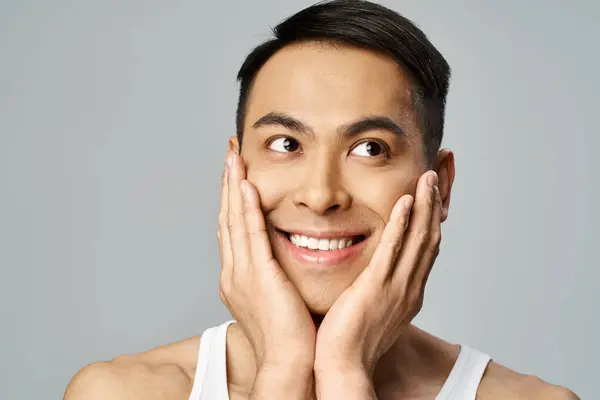 Hombre asiático guapo con una sonrisa serena, tocándose suavemente la cara en una rutina calmante para el cuidado de la piel en un estudio gris. - foto de stock