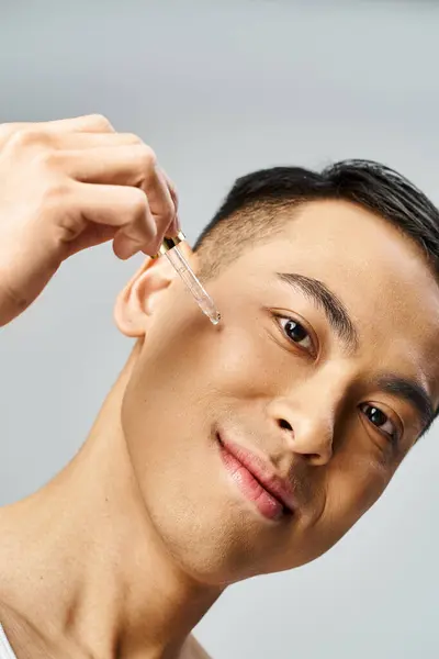 Азиатский мужчина улыбается и использует сыворотку в серой студии. — стоковое фото