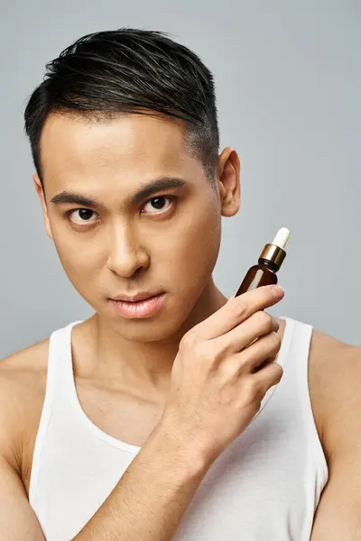 Beau asiatique tenant une bouteille de sérum dans un studio gris, se concentrant sur la routine des soins de la peau et les soins personnels. — Photo de stock
