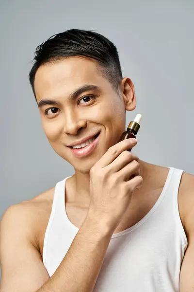 Счастливый азиат деликатно держит сыворотку в руке в серой студии, демонстрируя свою рутину с кожей. — стоковое фото