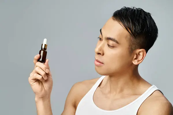 Un hombre asiático guapo en un estudio gris delicadamente sostiene una botella de aceites esenciales, que encarna la relajación y el autocuidado. - foto de stock