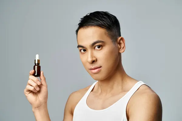 Un hombre asiático guapo sostiene una botella de aceite en un estudio gris, mostrando su rutina de cuidado de la piel con precisión y estilo. - foto de stock