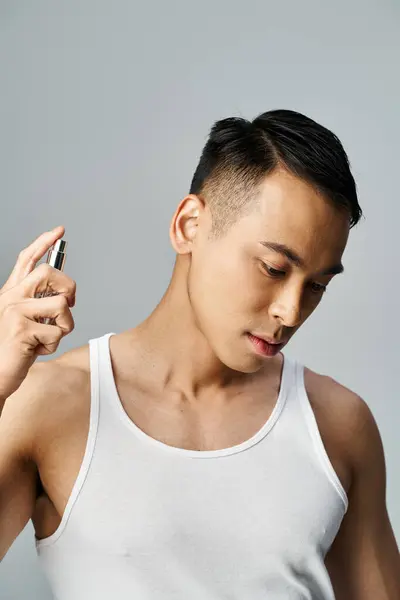 Un bel homme asiatique dans un débardeur blanc vaporisant du parfum dans un studio gris, se concentrant sur sa routine de toilettage. — Photo de stock