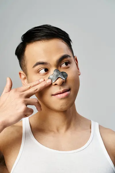 A handsome Asian man holding a piece of nose patch, showcasing his beauty and skincare routine in a grey studio. — Stock Photo
