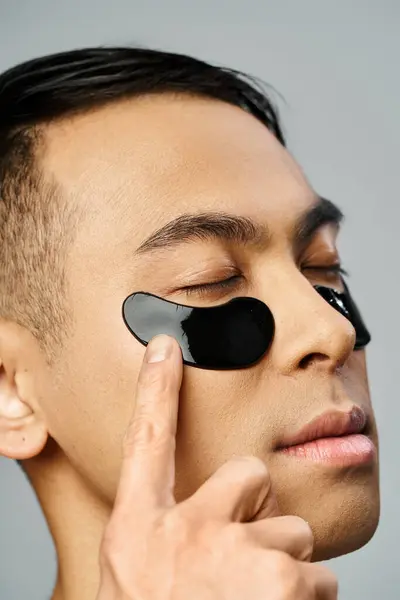 Asiatique homme avec un bandeau noir frappant améliorer sa peau lors d'une routine de beauté dans un studio gris. — Photo de stock