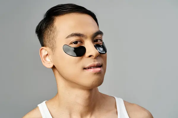 Hombre asiático guapo en un estudio gris usando parches para los ojos por su belleza y rutina de cuidado de la piel. - foto de stock