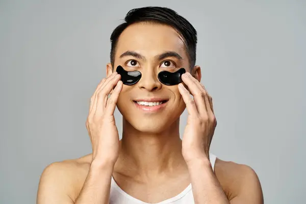 Азиатский мужчина держит два черных круга над глазами в красоте и уходе за кожей в серой студии. — стоковое фото