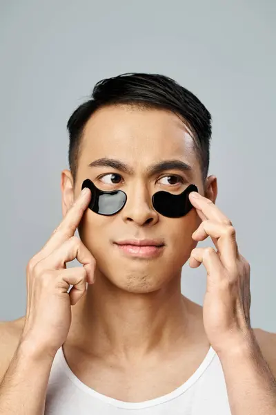 Handsome Asian man holding two black circles over his eyes in a beauty and skincare routine in a grey studio. — Stock Photo
