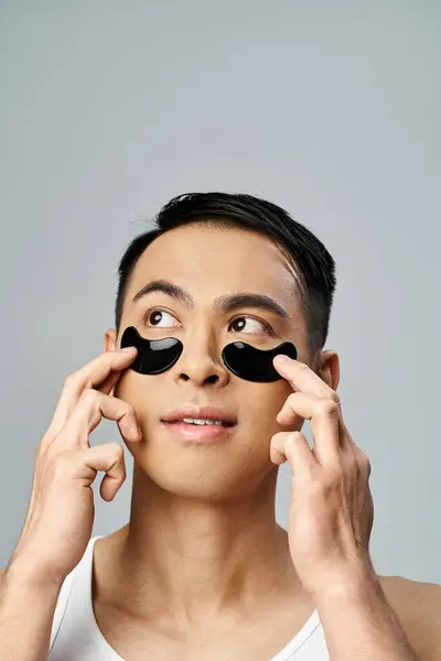 Handsome Asian man with under eye patches engaging in a beauty and skin care routine in a grey studio. — Stock Photo
