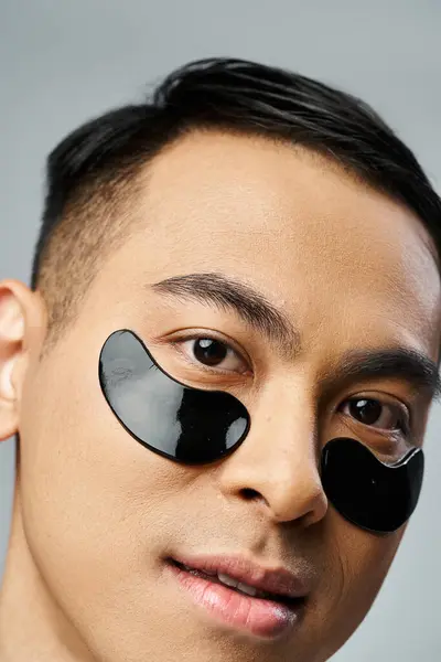 Hombre asiático guapo con parches debajo de los ojos durante la rutina de belleza y cuidado de la piel en estudio gris. - foto de stock