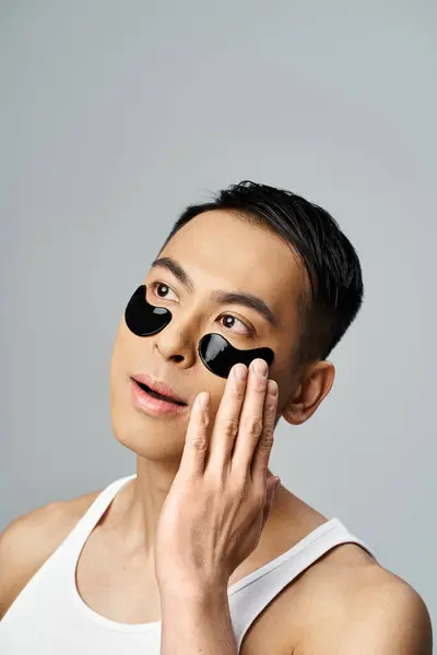 Handsome Asian man with black eye patches, in a beauty and skin care routine, in a grey studio setting. — Stock Photo